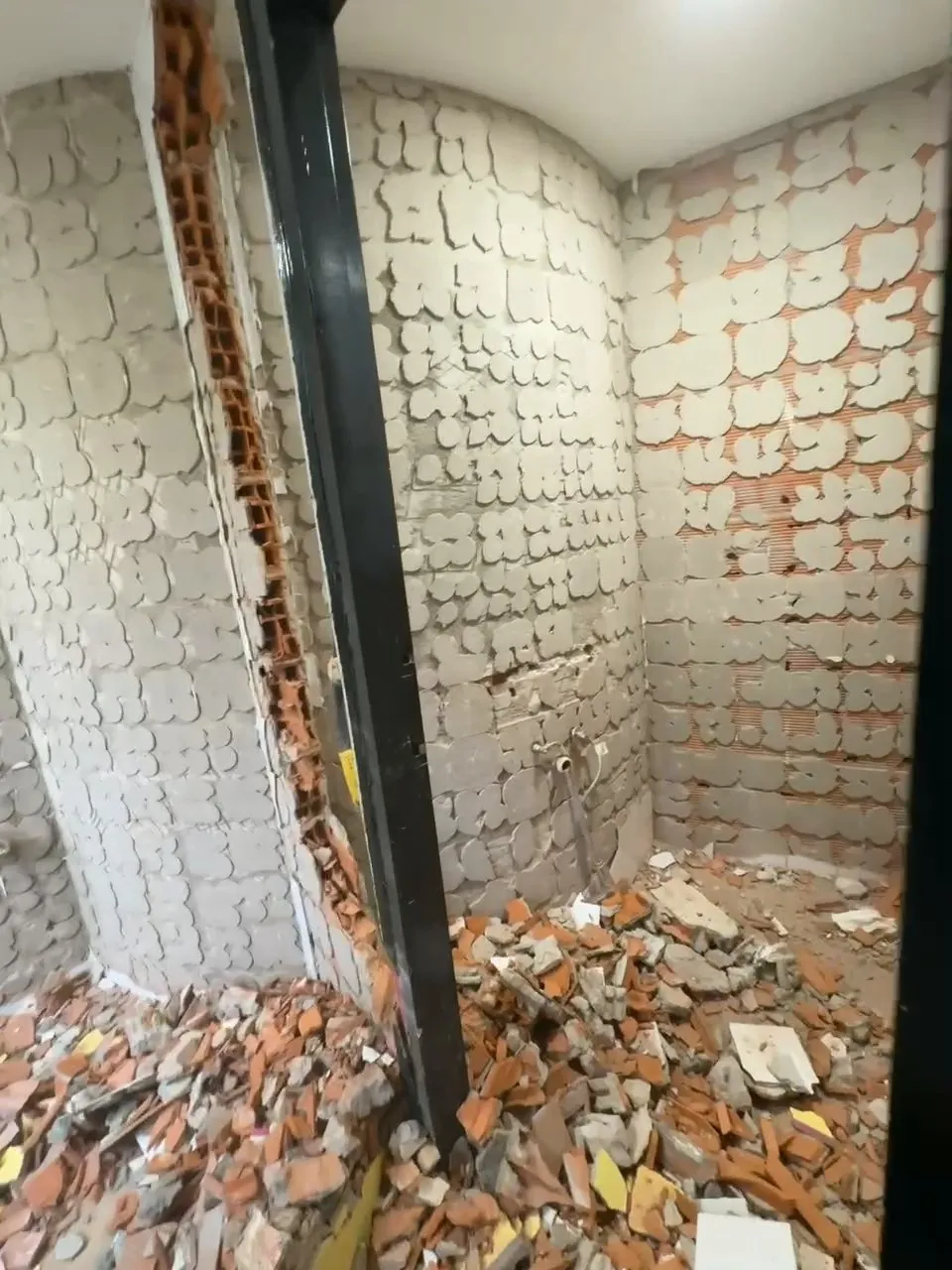 Interior d’un bany en enderroc: parets sense enrajolat, amb restes d’adhesiu i maó foradat a la vista, un perfil negre al centre i un munt de runa de maó i ceràmica pel terra