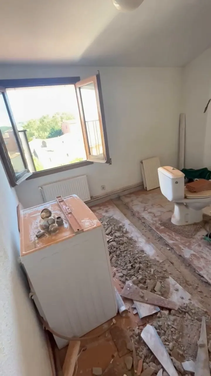 Estancia en obra con el suelo levantado por franjas y escombros, un inodoro con cisterna al fondo a la derecha, un electrodoméstico blanco apartado y una ventana abierta con vistas a tejados y árboles