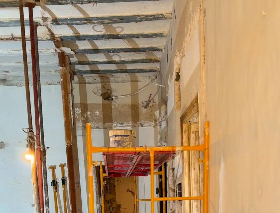 Estancia en obra con el techo de vigas al descubierto, puntales metálicos que llegan hasta él, una lámpara de obra encendida y un andamio amarillo con una plataforma roja y un cubo encima
