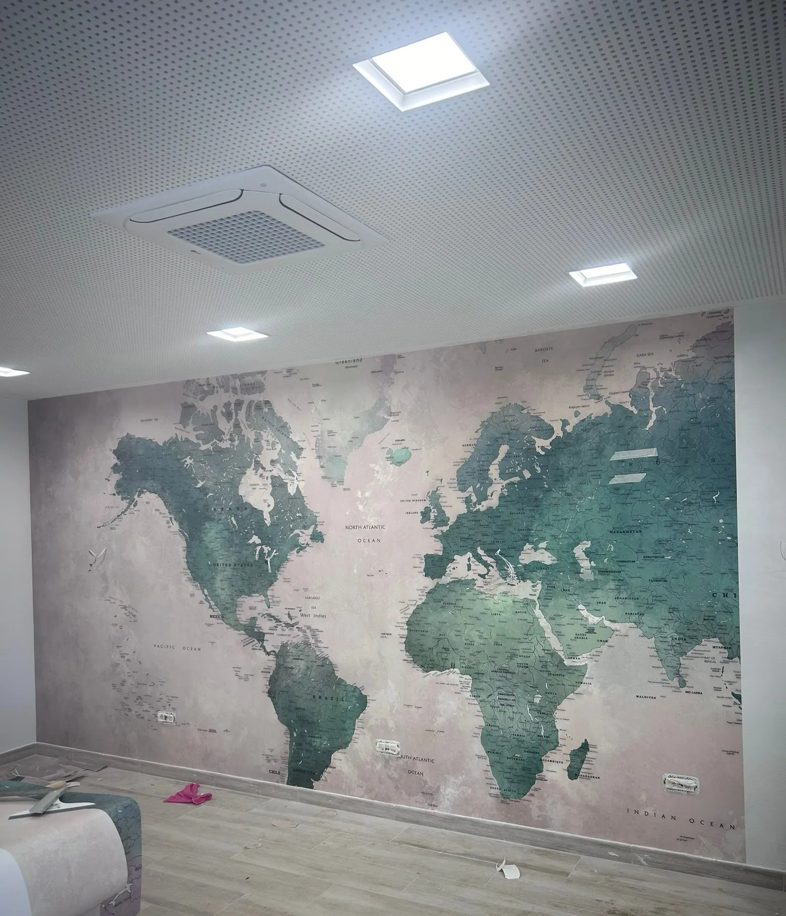 Paret coberta amb un mural de mapamundi durant la col·locació, amb un rotlle de paper, eines i restes a terra; fals sostre perforat amb focus i climatitzador