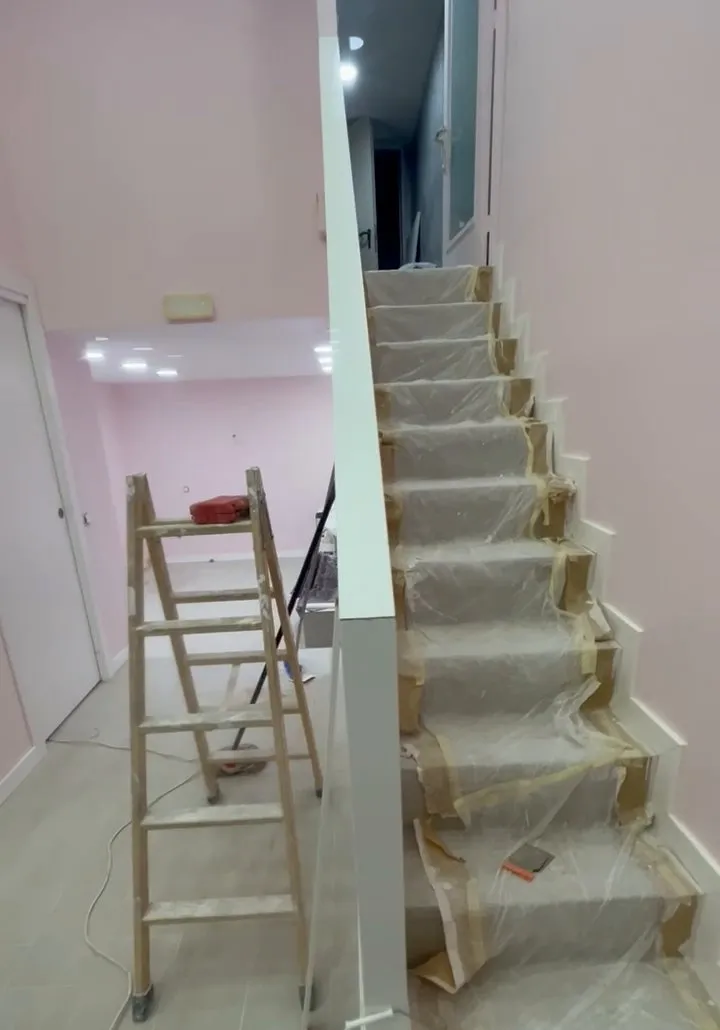 Escala amb els graons coberts amb plàstic i cinta de pintor, al costat d’una paret rosa pàl·lid i un muret blanc; a l’esquerra, una escala de mà de fusta amb restes de pintura i, al fons, una sala amb focus al sostre