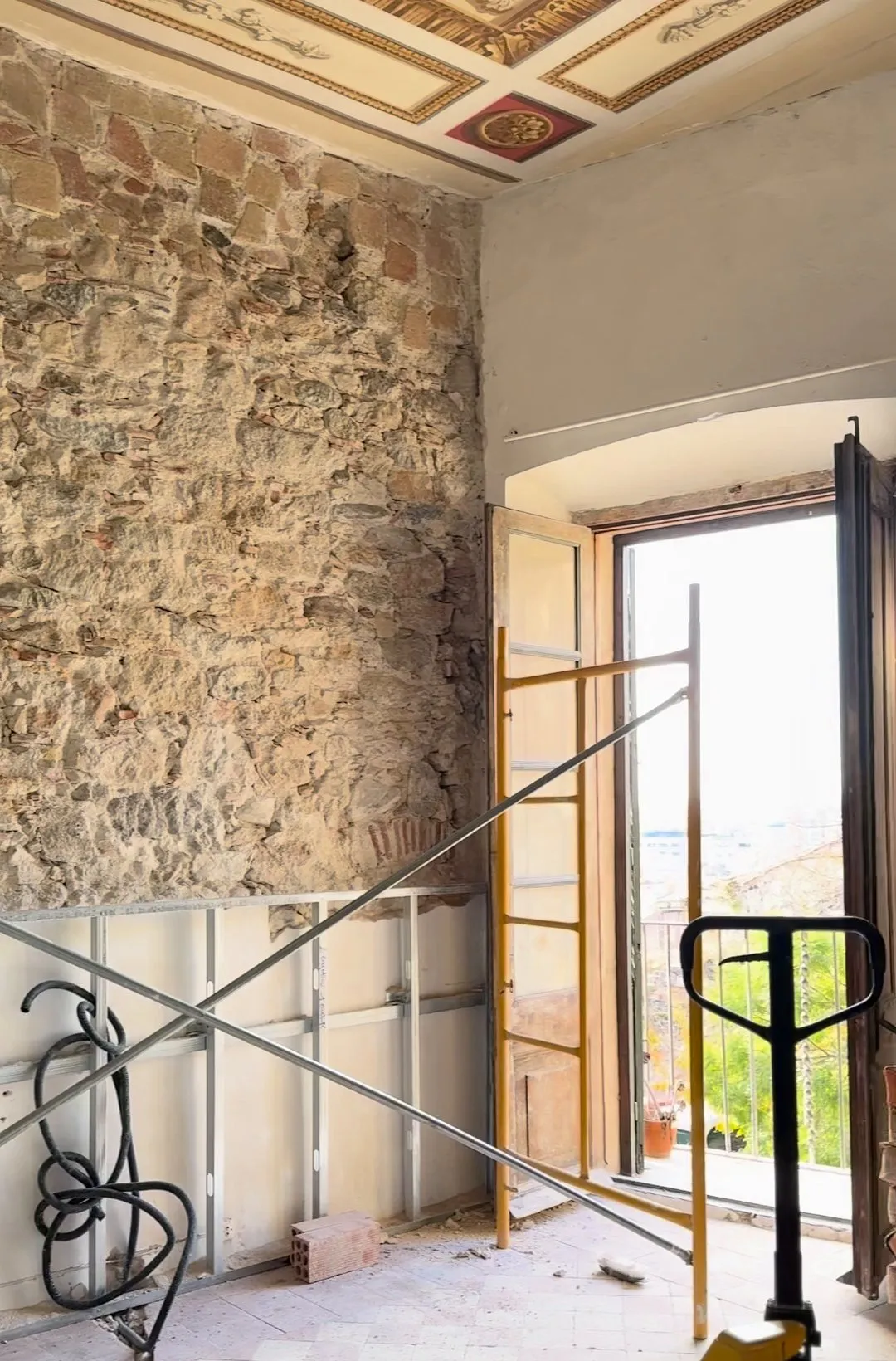 Estancia en obra con un muro de piedra vista, perfiles metálicos montados en la parte baja de la pared, un andamio amarillo, una balconera abierta y el techo con molduras pintadas