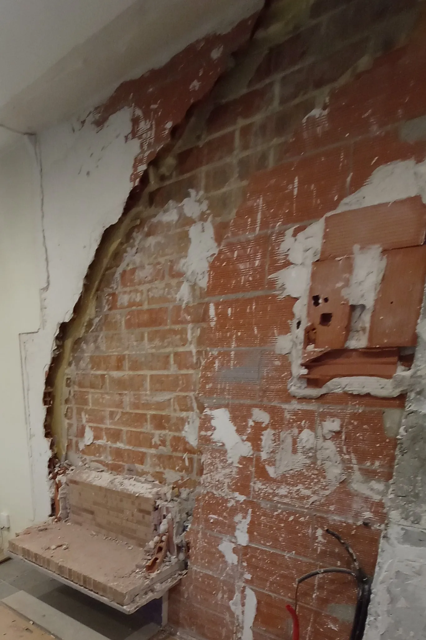 Pared de ladrillo a la vista, con restos de yeso y el contorno curvo del revestimiento arrancado; abajo a la izquierda queda la base de ladrillo del hogar y a la derecha cuelgan unos cables