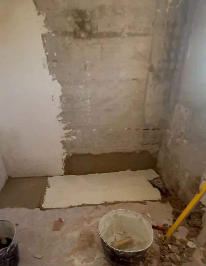 Rincón de una estancia en obra con la pared sin revestir y restos de acabado, una franja de mortero a ras de suelo con una zona blanca extendida encima, un cubo con una herramienta de mano y un nivel amarillo apoyado en la pared