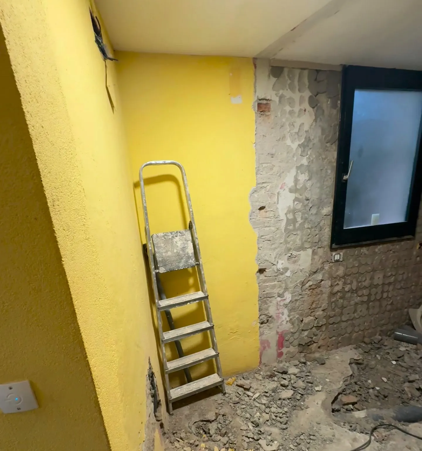 Bany en obres amb la paret de la finestra picada, amb restes d’adhesiu on hi havia l’enrajolat, una finestra de marc negre, una escala metàl·lica recolzada en una paret groga i runa pel terra