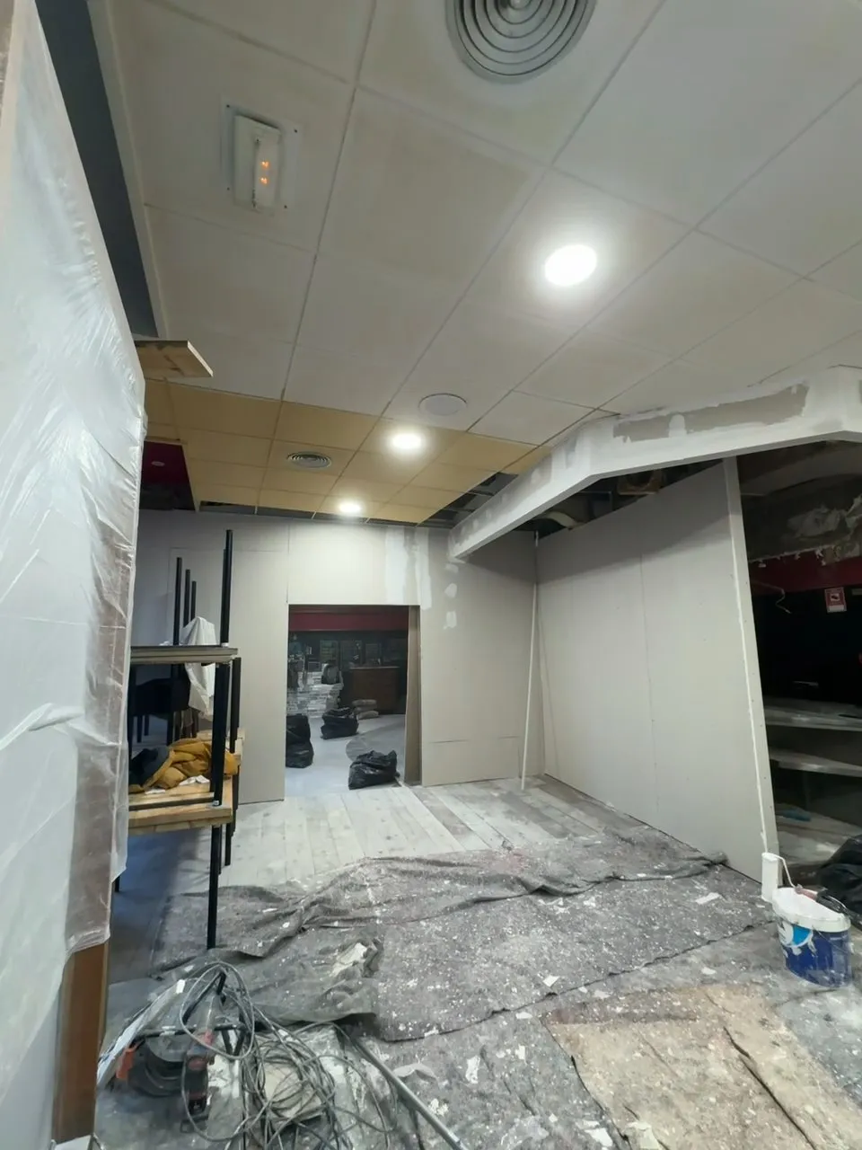 Vestíbul en obres amb envans nous de placa de guix laminat sense pintar, un tram de sostre rebaixat amb les juntes tractades, fals sostre de plaques amb focus encastats i el terra cobert amb proteccions, cables i una galleda de pintura amb corró
