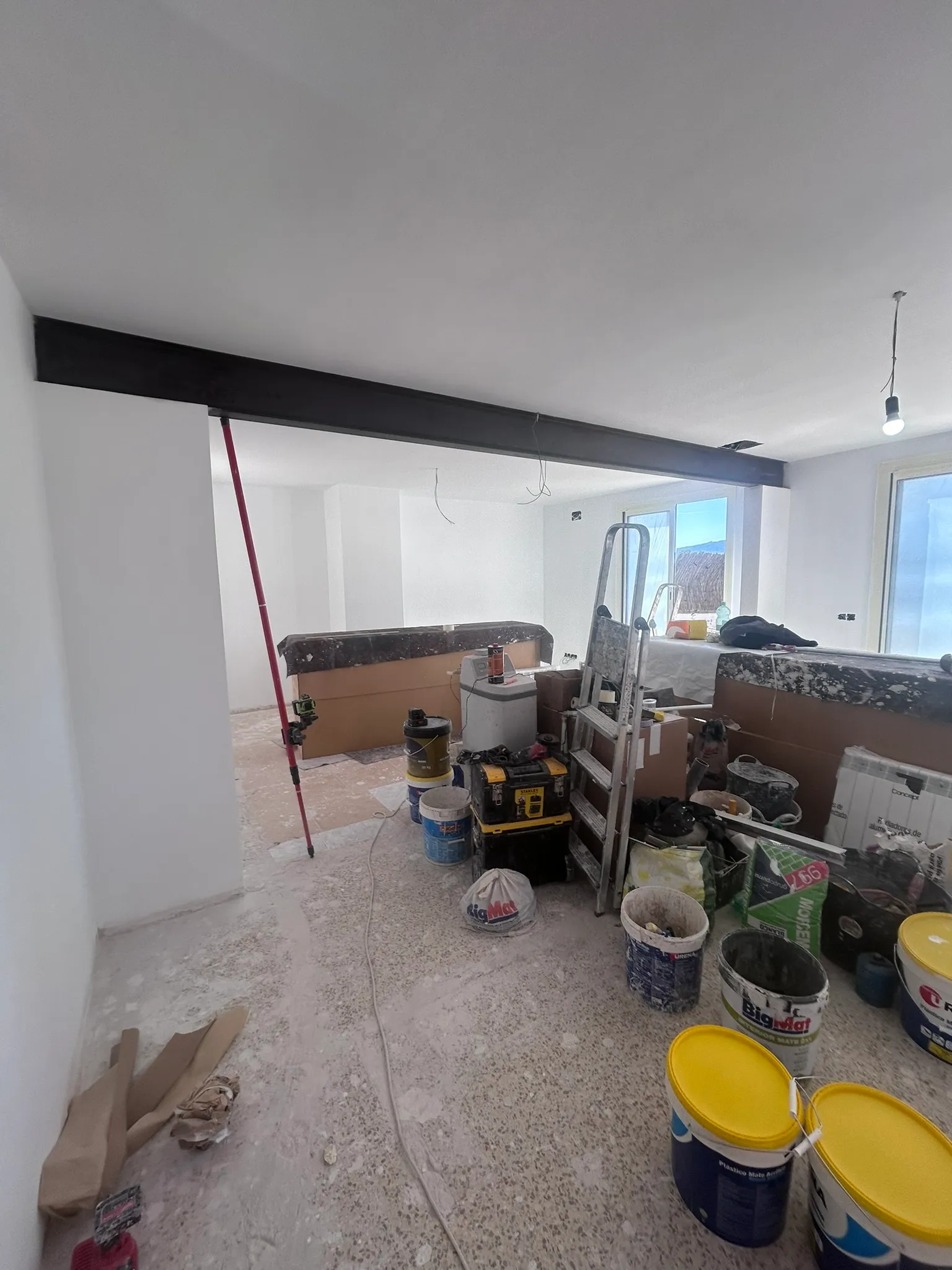Viga de acero instalada en un vano abierto entre dos estancias, con puntal, escalera y materiales de obra.