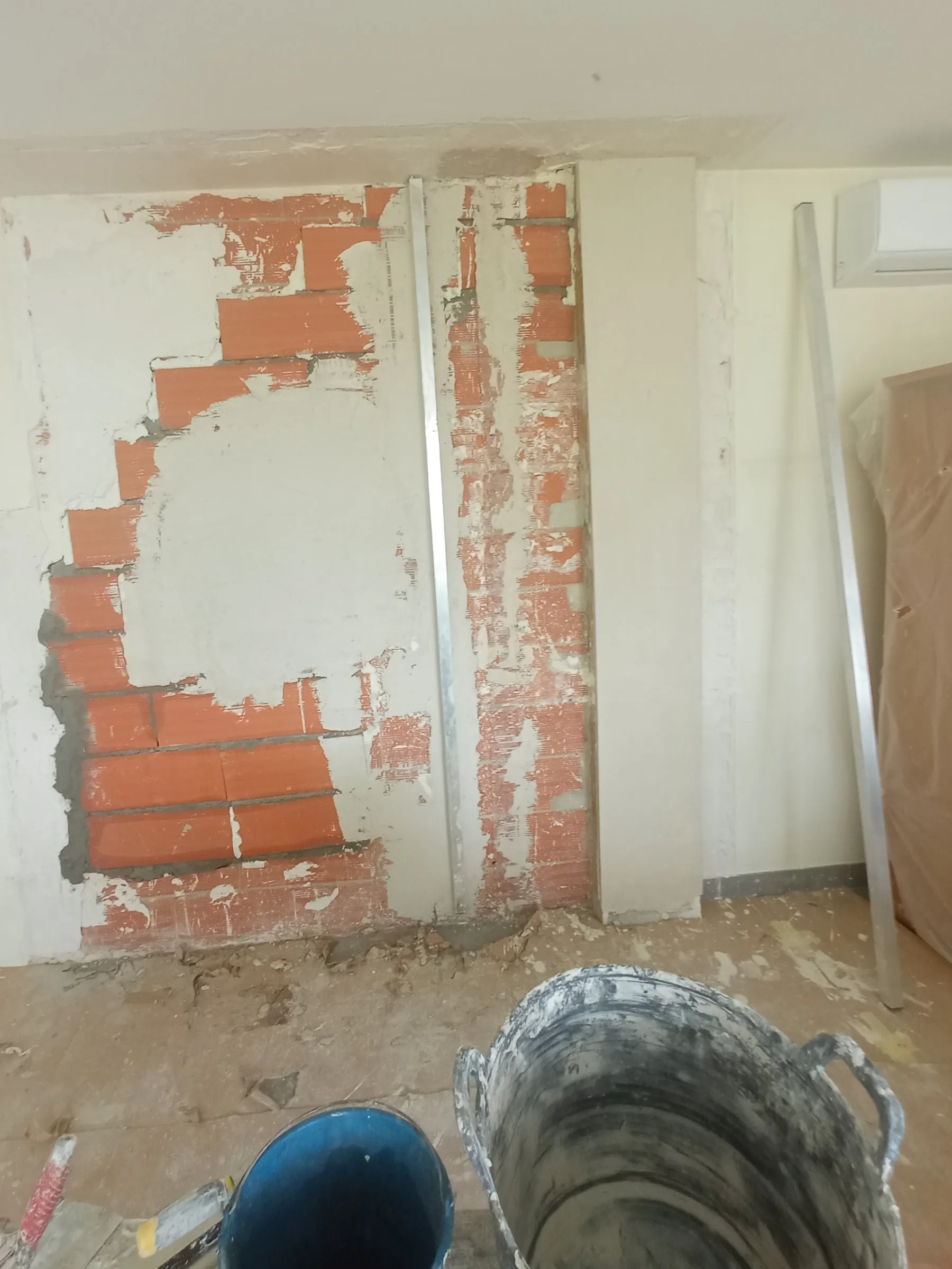 Pared a medio enyesar: parches de yeso sobre el ladrillo, una franja de ladrillo a la vista, un perfil de aluminio vertical y, en primer plano, un cubo azul y un recipiente grande de goma con restos de mezcla