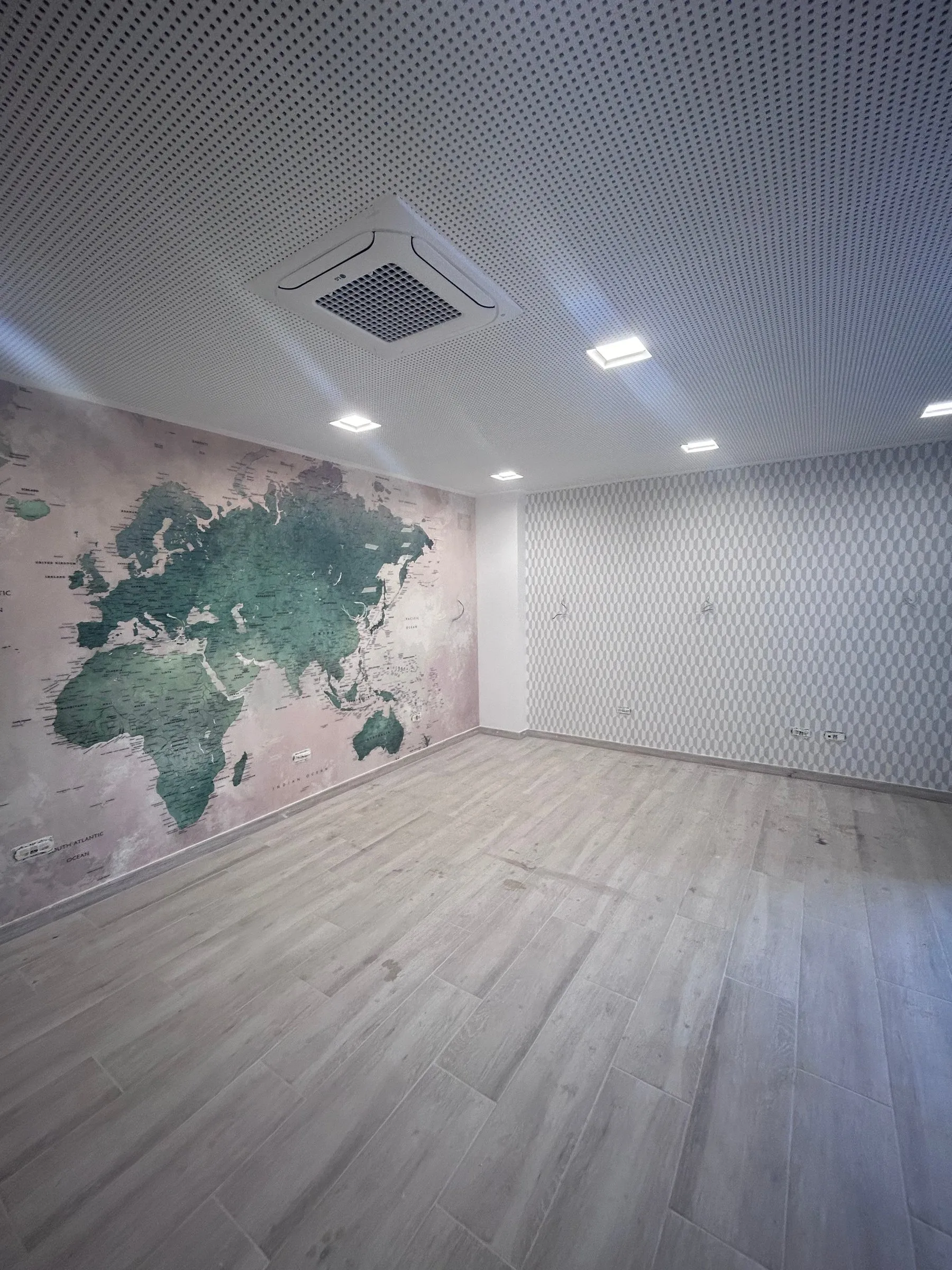 Sala sense moblar amb un mural de mapamundi en una paret i paper pintat geomètric a l’altra, terra d’aspecte de fusta clara i fals sostre perforat amb focus