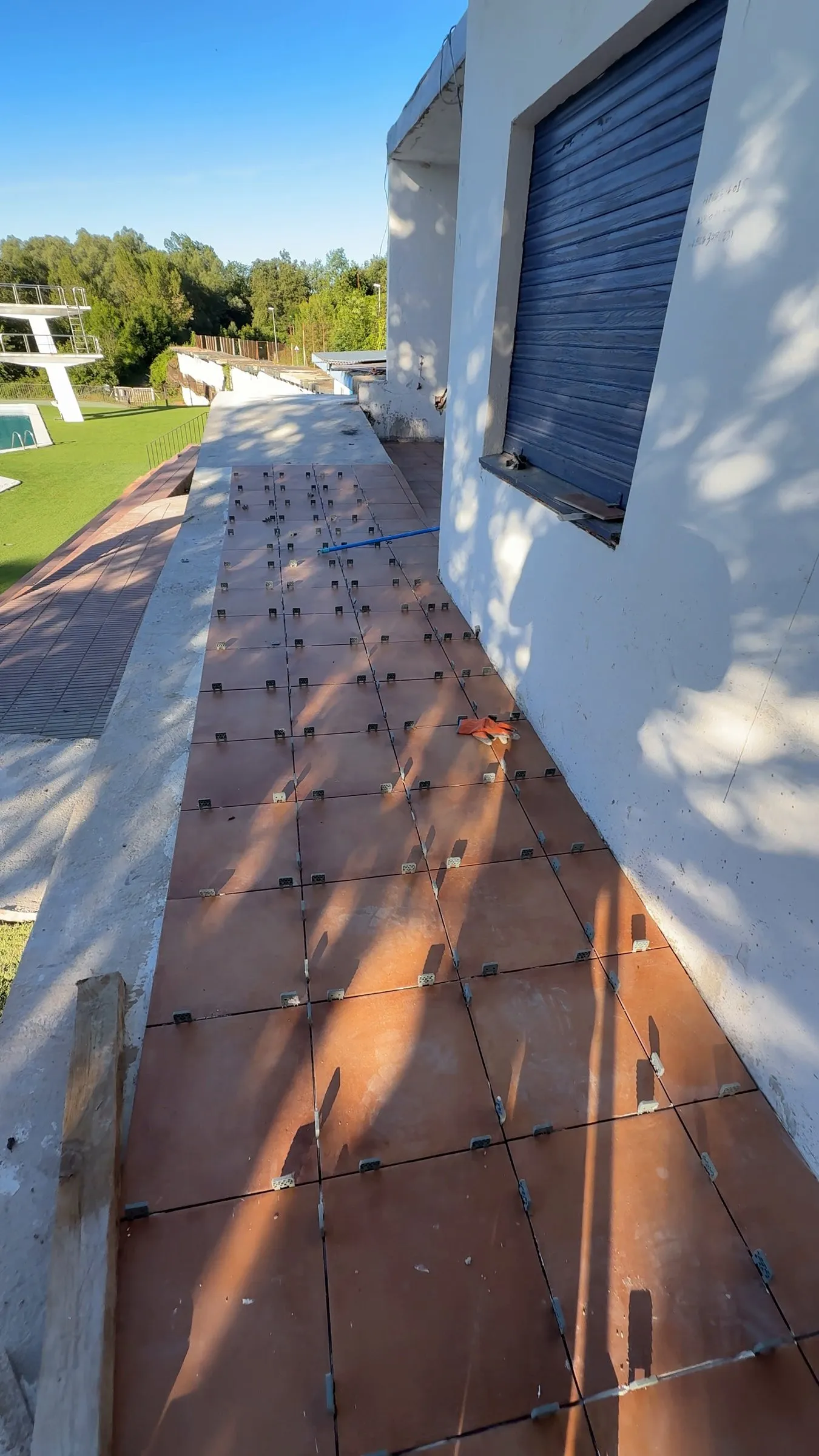 Baldosas cerámicas de color terracota colocadas sobre una base de hormigón, con separadores de nivelación entre las piezas, junto a una pared blanca con persiana azul y, al fondo, la piscina y el trampolín