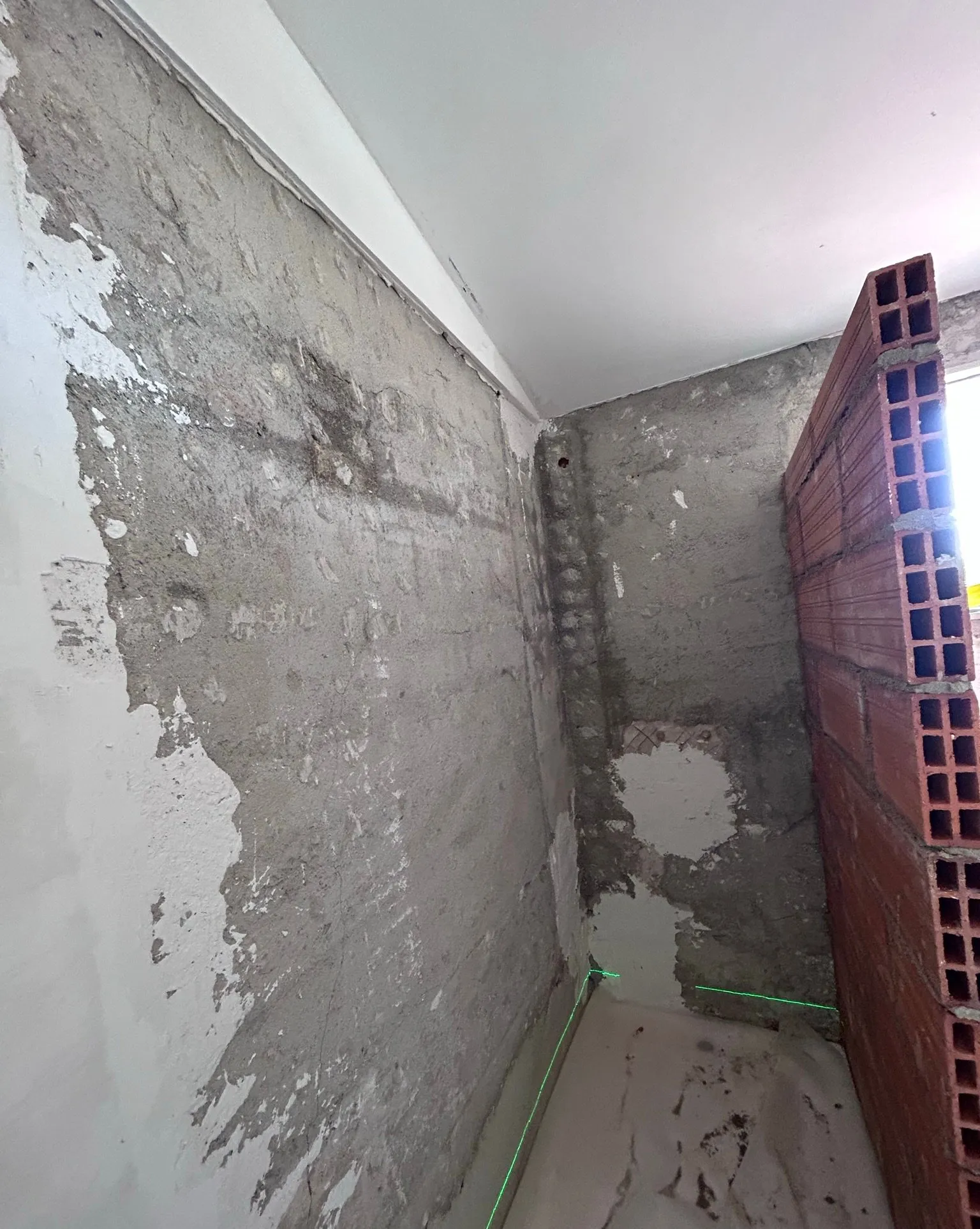 Rincón de una estancia en obra con las paredes sin revestir, un tabique nuevo de ladrillo hueco a la derecha y, abajo, una base blanca con líneas verdes de láser de nivelación