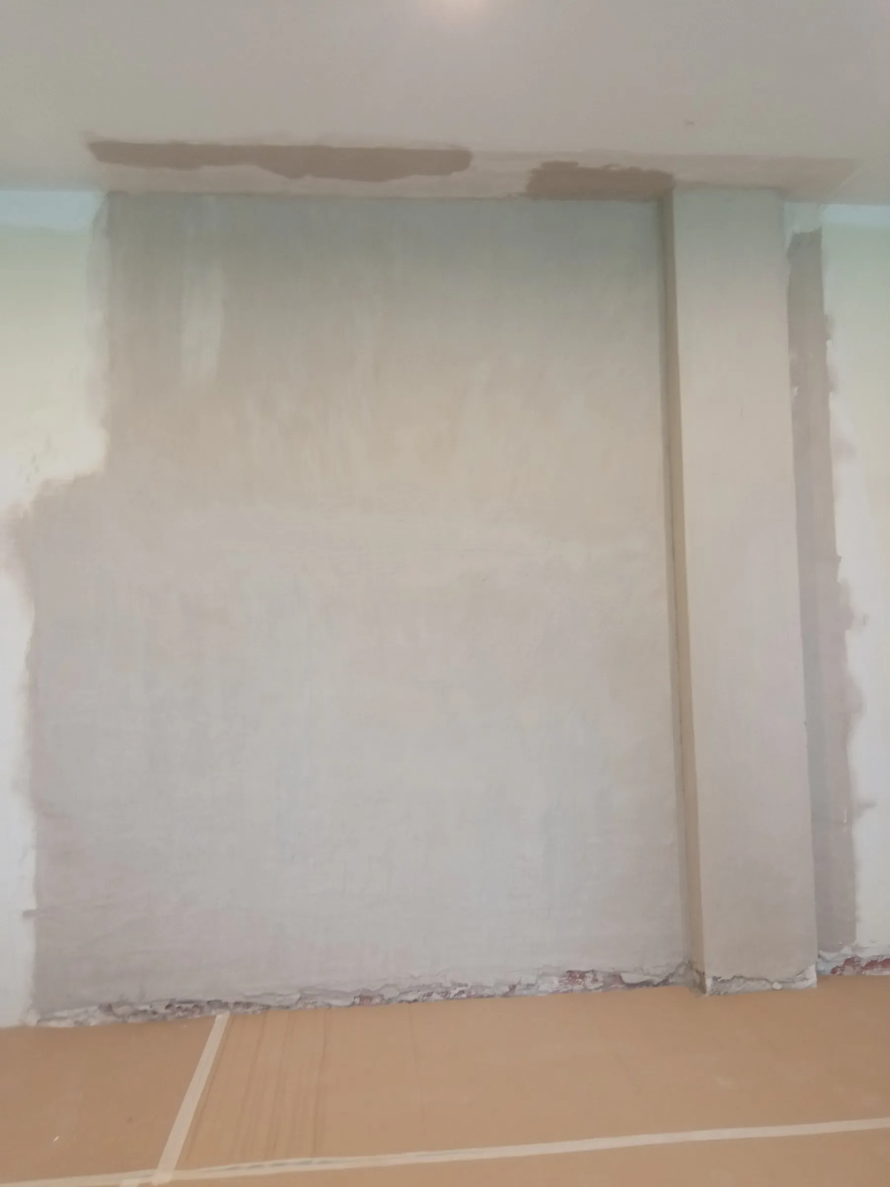 Pared enyesada de color gris claro, con zonas de otro tono en el borde izquierdo y en la franja del techo, un resalte vertical a la derecha y el suelo protegido con cartón y cinta