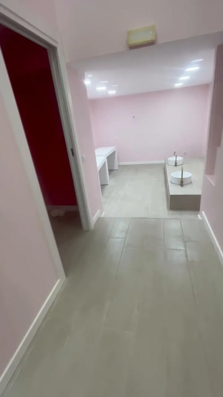Passadís que dona a la sala del local, amb parets rosa pàl·lid, terra de lames gris clar i sòcol blanc; al fons, dues taules blanques i una plataforma elevada amb dos lavabos rodons blancs amb aixeta daurada, sota focus encastats, i a l’esquerra, la porta oberta d’un bany pintat de fúcsia