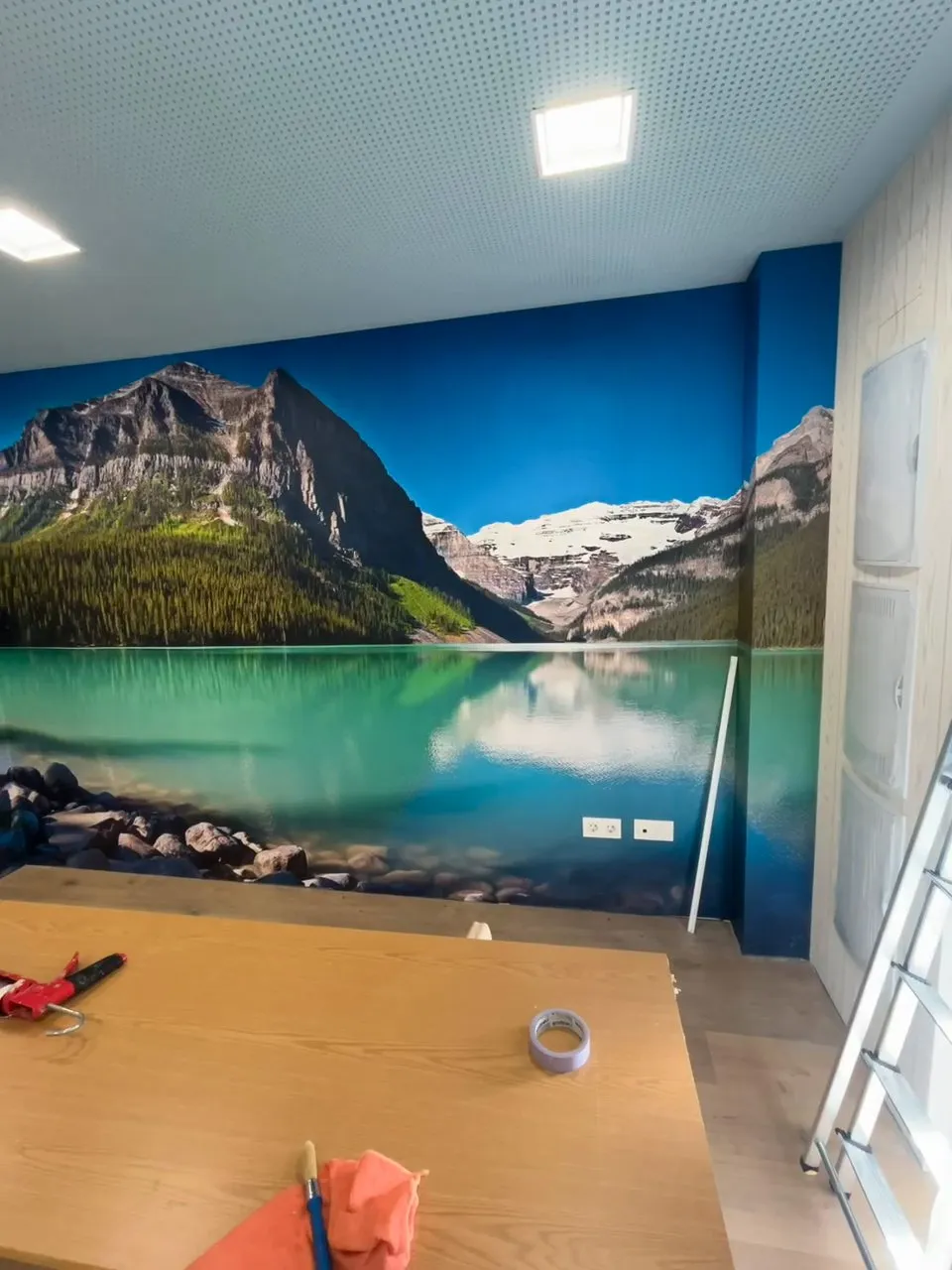 Sala en obres amb un mural d’un llac de muntanya ja col·locat, terra d’aspecte de fusta, fals sostre perforat amb focus i una taula de treball amb eines