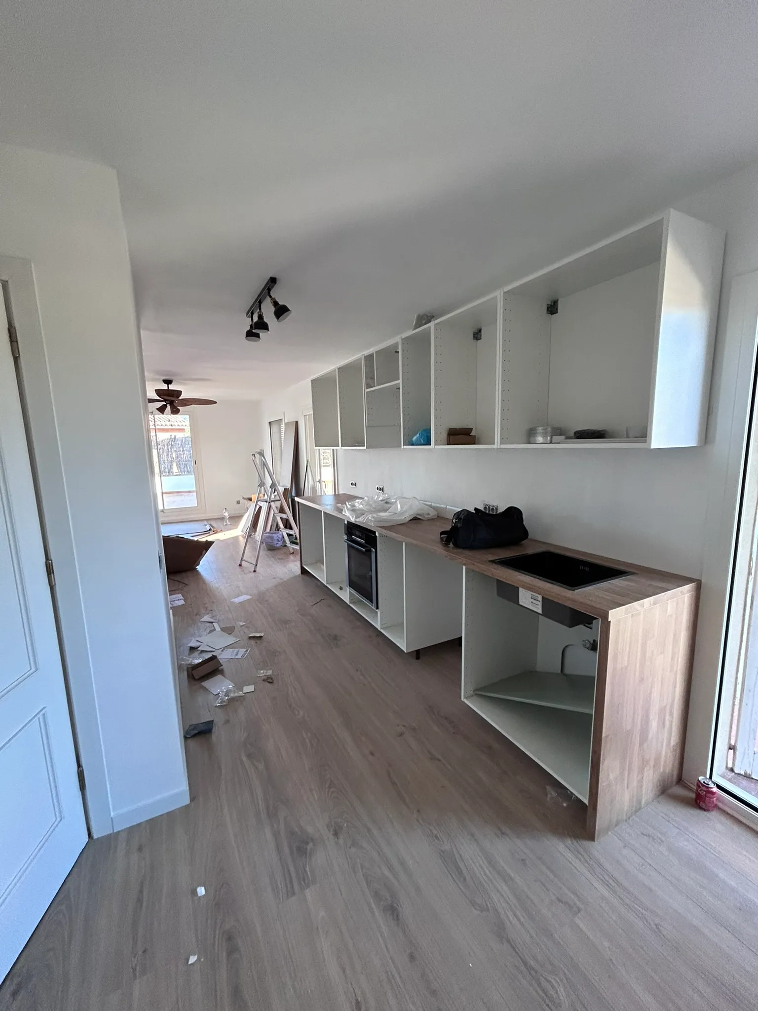 Muebles de cocina montados sin frentes, con la encimera con acabado de madera y el horno ya colocados; al fondo, sin tabique de por medio, se ve el salón con el ventilador de techo