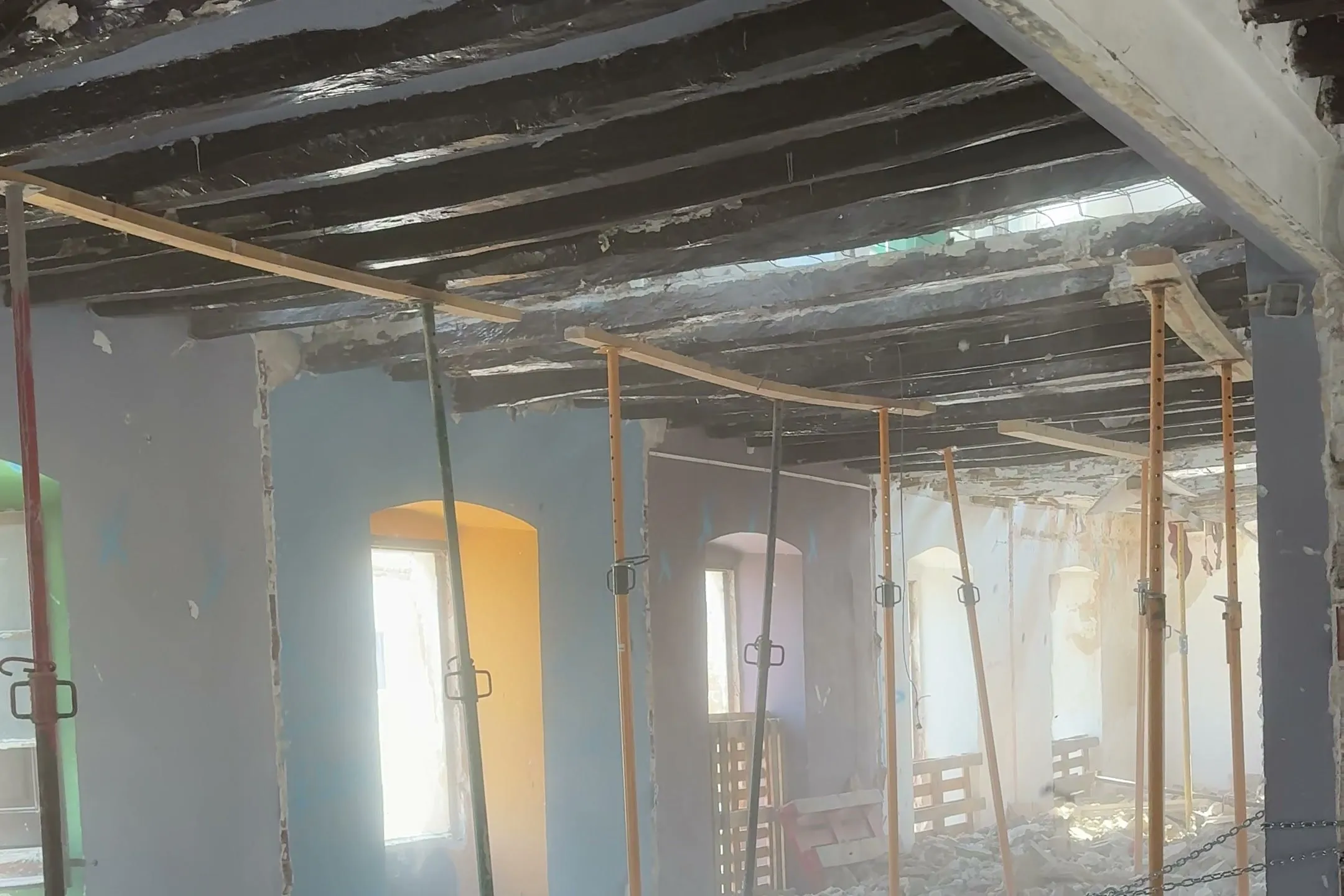 Sala de una vivienda en rehabilitación en Banyoles: puntales metálicos, techo de vigas de madera y ventanas en fila.