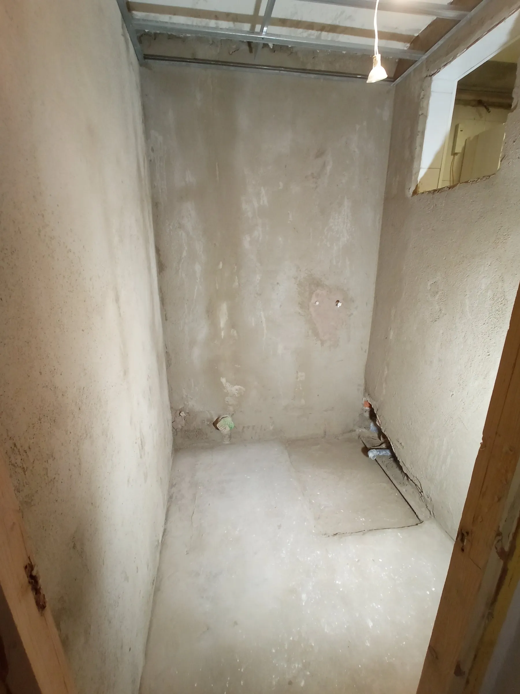 Cuarto de baño en bruto: paredes y suelo sin revestir, perfiles metálicos del falso techo, una bombilla colgada de un cable y un hueco en la pared