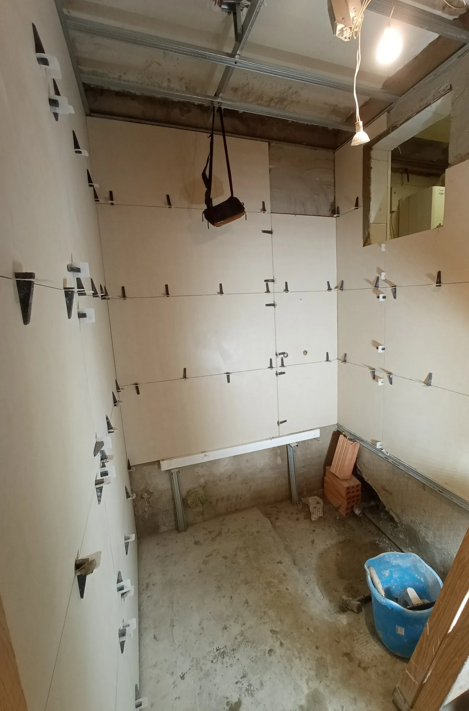 Colocación de baldosas grandes de color crema en las paredes del baño, con clips y cuñas de nivelación y un cordel de guía; el suelo sigue en bruto