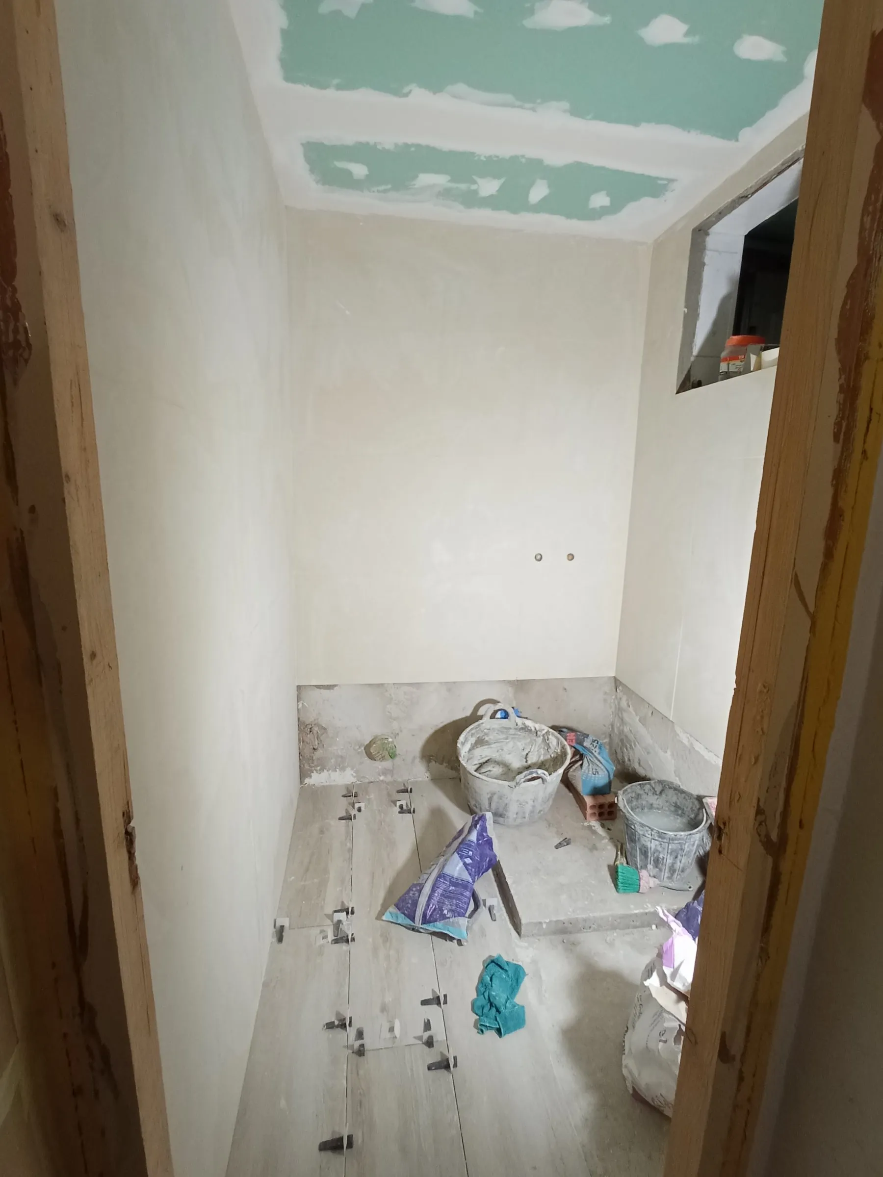 Baño en obra con las paredes alicatadas de color crema, falso techo de placas verdes con las juntas tratadas y el suelo de baldosa de aspecto de madera a medio colocar