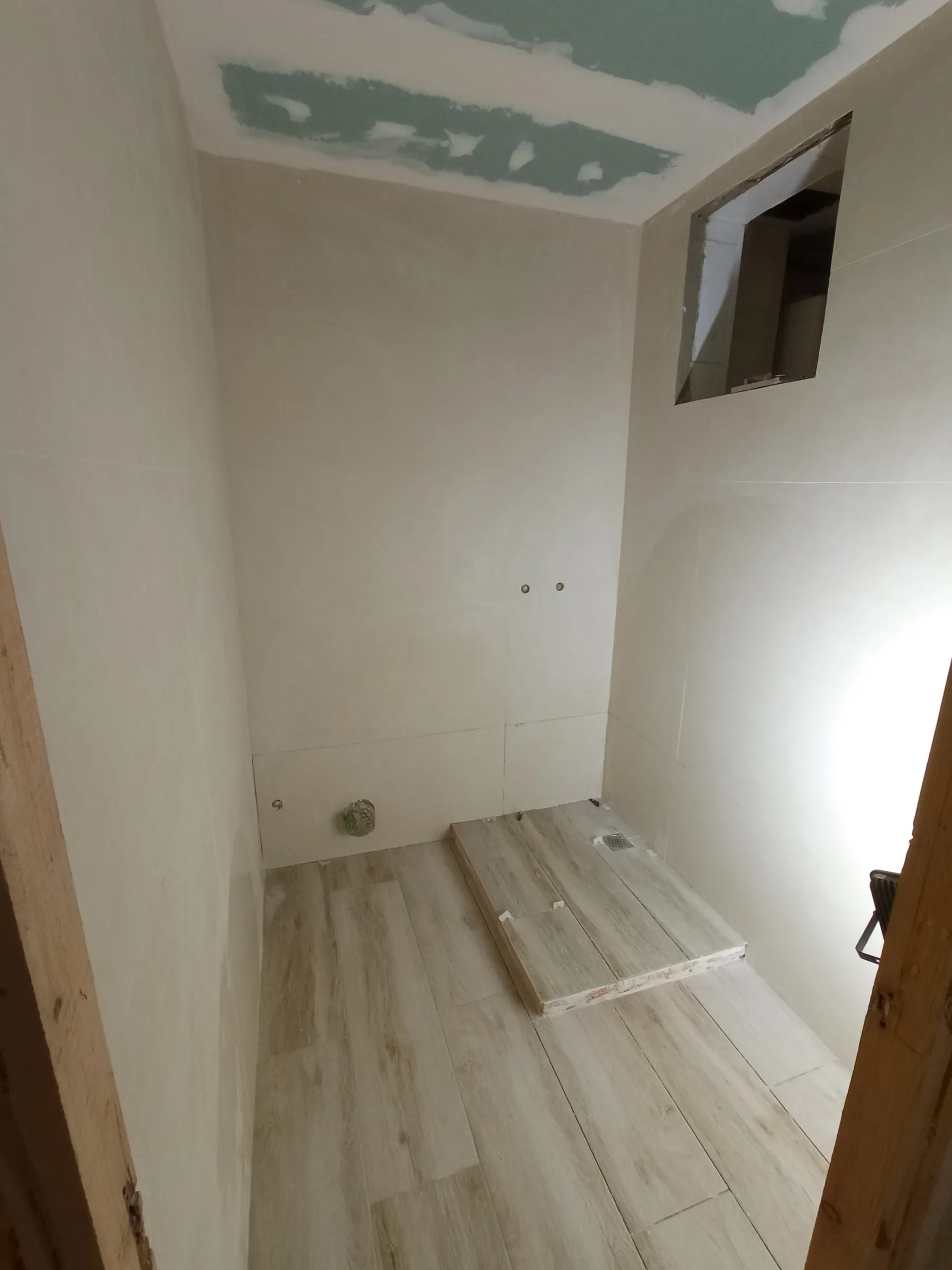 Baño en obra con alicatado y suelo colocados: baldosa de aspecto de madera y, a la derecha, una base elevada con desagüe revestida con la misma baldosa; todavía sin sanitarios